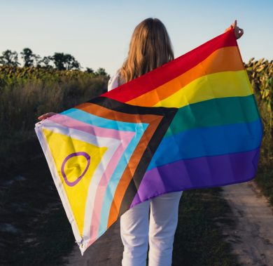Symbol of LGBTQ pride month. Young woman showing Rainbow LGBTQIA flag waving in wind made from silk material on field background. Equal rights. Peace and freedom concept